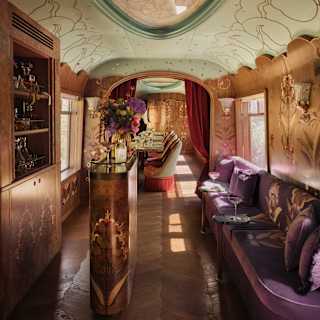 Luxurious vintage train carriage interior with ornate wood panelling, velvet seating, floral arrangements, decorative ceiling panels, and soft ambient lighting. A long table is visible further down the carriage.