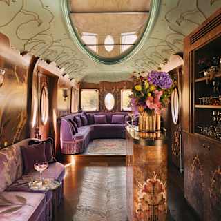 Luxurious vintage train carriage interior with ornate floral decor, plush purple seating, an oval ceiling mirror, flowers on a gold stand, and a built-in bar, all bathed in warm, elegant lighting.