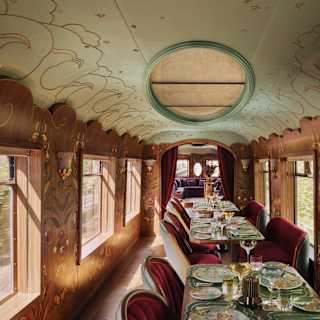 Luxurious vintage train dining carriage with ornate decor, set tables with glassware and plates, red velvet chairs, large windows, and elegant lighting, creating an inviting and nostalgic atmosphere.