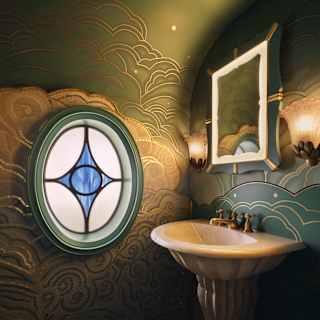 A small bathroom with ornate green and gold patterned walls, a pedestal basin, a square mirror with two wall lights, and a round stained glass window featuring a blue diamond shape.
