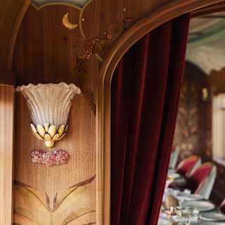Art Deco-style wall sconce with a lotus-shaped glass shade and floral designs on wood panelling, next to a deep red curtain that opens into a dining area set with plates and glassware.