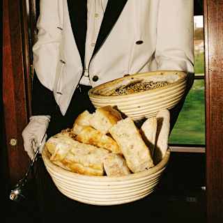 A waiter in a black and white blazer with gold shoulder cords carries two baskets of cut bread on one arm, seen in close-up.