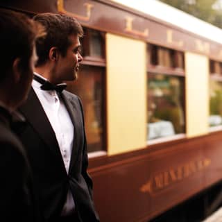 2 men in dinner jackets stand on the train platform and look at the length of the cream and burgundy British Pullman train