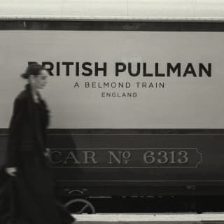 Black and white soft-focus image of a woman in a long coat walking on the platform past a car with the British Pullman brand.