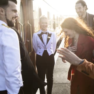 A uniformed male and female member of staff laugh with a happy woman as she and two male friends prepare to board the train.