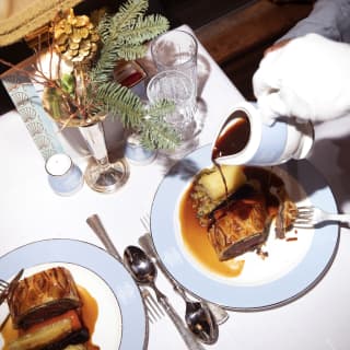 A waiter's gloved hand pours gravy over crisp Beef Wellington as a couple indulge in seasonal fine dining, seen from above.