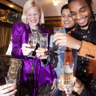 A group of friends dressed up for the evening in bright, luxurious clothes, chink glasses as they party in the Zena carriage.