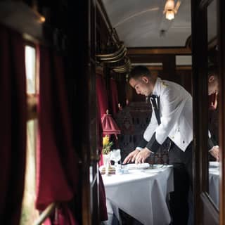 Red velvet curtains frame the windows as a waiter, dressed in white jacket with gold epaulettes, sets the table for dinner