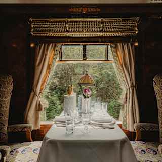 Looking at a table set for two and flanked by luxurious chairs, illuminated by the window and framed by tied-back curtains.