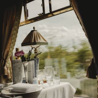 A green view rushes past the window of a dining table set for two with Champagne, bud vase and lamp, seen at eye-level.