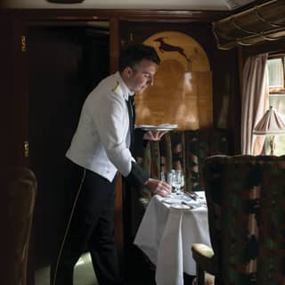A waiter sets a table in an art deco carriage. Divine marquetry on the wall behind him shows a gazelle leaping a gold hillock