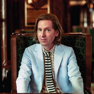 Wes Anderson smiles quizzically, dressed in a pale blue seersucker blazer, yellow and navy striped tie and yellow plaid shirt