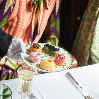 A waiter's white gloved hand sets a plate of mini pastries onto the table, including a pink macaron and raspberry mousse tarte