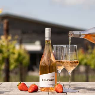 A bottle of Balfour Nannette's Rosé stands on a table among vines, as two glasses of the pale still blush wine are poured