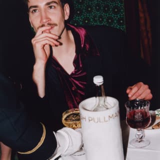 A waiter delivers a drink to a man with a moustache in a satin red top, at a table with food and a bottle in a wine sleeve.