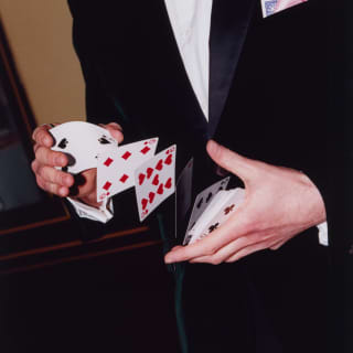 Playing cards are snapped in mid-air as a magician in a black suit with a gold bow-tie and pink handkerchief riffles a deck.