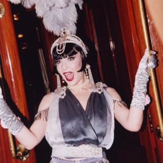 A woman with a dark bob, wearing a satin grey dress and gloves, and a tall feather fascinator, poses in the doorway of a carriage.