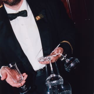 A male staff member holds a glass in one hand, and four upturned wineglasses, with the stems between his fingers, in the other.