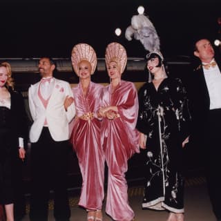 A line-up of five female performers in Art-Deco gowns and headdresses and three men in black, white and pin-stripe suits.