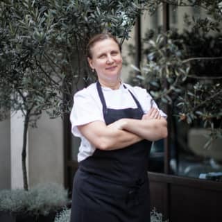 Michelin-starred chef, Angela Hartnett, stands with arms folded on a terrace surrounded by a grove of potted olive trees