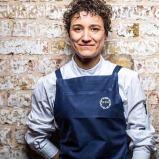 Michelin-starred chef, Nieves Barragán Mohacho, leans on a wall in her chef's whites and navy apron with the Sabor logo