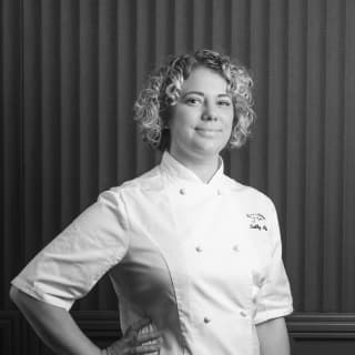 A black and white image of acclaimed British chef, Sally Abé, as she poses for the camera in chef's whites with her hand on hip
