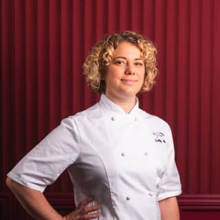 Acclaimed British chef, Sally Abé, poses for the camera in signature chef's whites with her hand on hip in front of a red wall