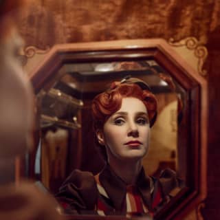 A red head with 1950s-style soft waves looks at herself in a mirror. She is dressed in striped blouse, buttoned at the neck