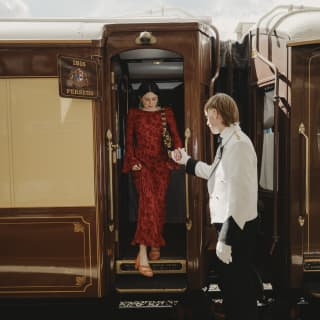 A male steward helps a female guest, wearing a luxurious red dress, step down from the train carriage on to a welcome carpet.