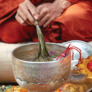 Close-up of an orange-robed monk dipping palm leaves in a silver bowl