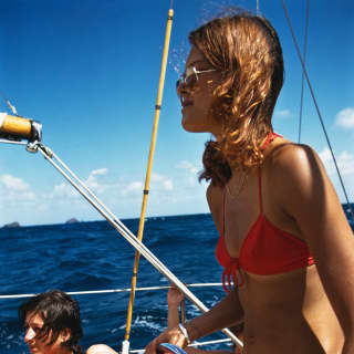 A tanned woman in a red bikini and sunglasses stands at the wheel of a sailing boat while friends relax behind.