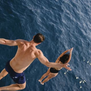 Action shot from above of a man leaping from a height following a woman forming a dive - both in mid-air with the sea below.