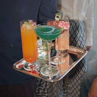 Close-up of a silver tray with five vibrant cocktails served in a diverse range of glass shapes, decorated with blossoms.