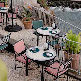 White-tiled tables with teal and pink chairs overlook rocks and sea through Brisa do Mar's terrace rails, seen from above.