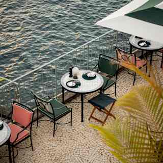 Looking down on a white-tiled round table for two with teal chairs on a mosaiced terrace overlooking sea in evening sun.
