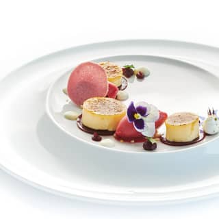 A white dessert plate with a curve of crème brûlées, tamarillo sorbet quenelles, chocolate discs and white chocolate drops.