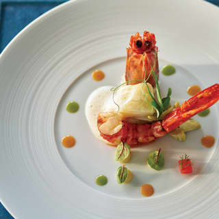 A prawn served in a white foam on a white plate, decorated with pea shoots, leaves and dots and cubes of orange and green.