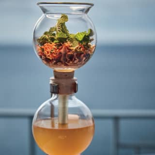 A candle-warmed coffee siphon containing glass chambers of botanicals and consommé on a restaurant table overlooking the sea.