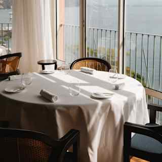 A round window table with four chairs, water glasses and rolled napkins catches the sun and sea views at William Restaurant.