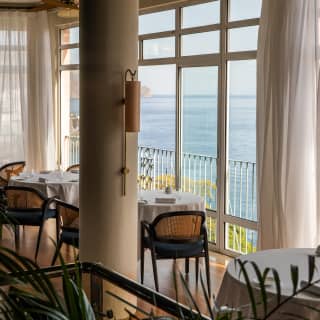 Dining tables set beside a drape-framed wall of windows soak up sea views, seen from behind a pillar in William Restaurant.