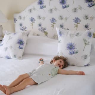 A smiling young child in short dungarees lies on their back with arms spread on a fresh white bed with floral cushions.