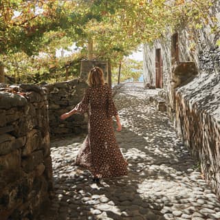 From behind, a woman in a long, floaty, orange and black dress walks in the dappled shade of a garden path with a sea vista.