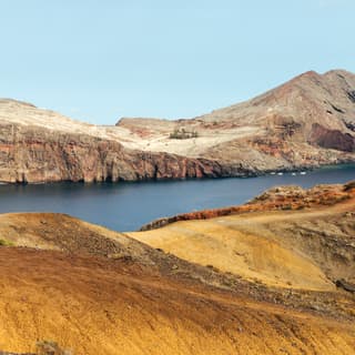 View over a rocky ridge with gold grasses to a deep blue inlet where four small boats are anchored by Ponta de São Lourenço.