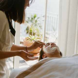 A spa therapist uses a brush to apply a white clay mask to a guest's face, with a glimpse of the ocean view balcony behind.