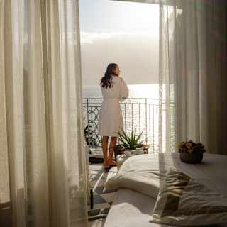 A woman in a robe leans on balcony railings gazing at the ocean, seen from the spa interior through an open door and drapes.
