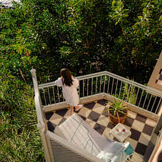 A woman in a white robe stands on a check-tiled balcony with lounge chair and table, gazing at the sea, seen from above.