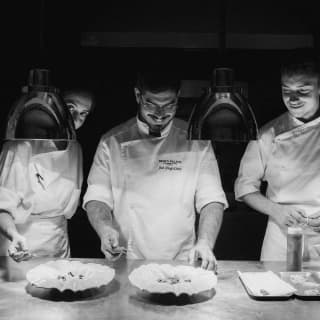 Behind a row of pendant heat lamps, Chef Costa and two chefs transfer garnish from trays to plates, seen in black and white.
