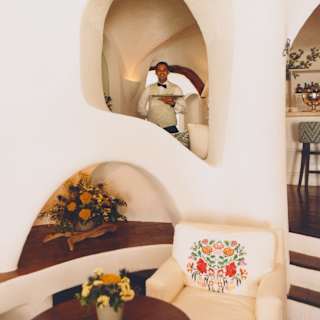 A smiling waiter, holding a tray, is glimpsed in a wall arch in a view of Bar Ginepro, capturing alcoves, armchair and table.