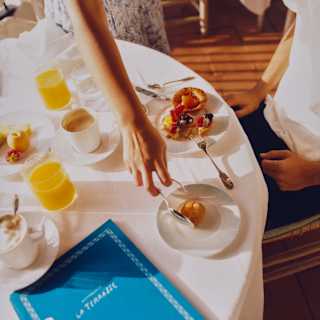 A waitress places a roll on the plate of a guest enjoying breakfast with coffee and juice at La Terrasse, seen from above.