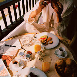 A guest enjoys a balcony breakfast at a sunny table with coffee, orange juice, toast, pastries and fruit, seen from above.